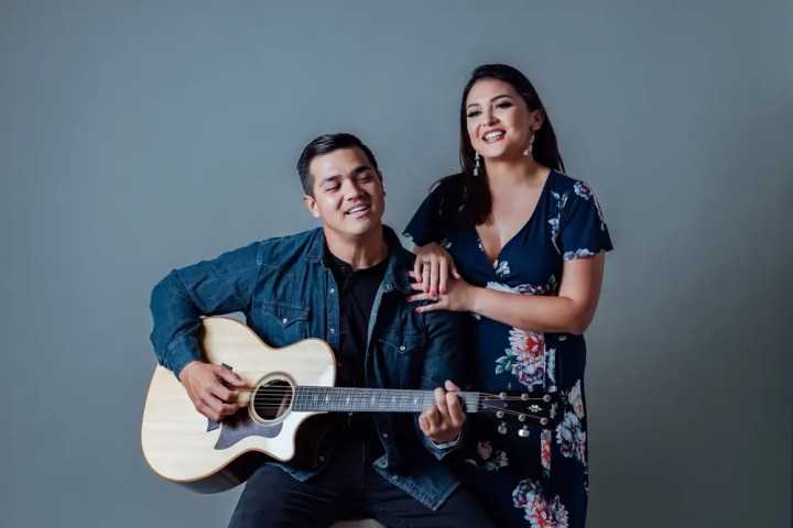 Man playing guitar with woman smiling, standing beside him in floral dress.