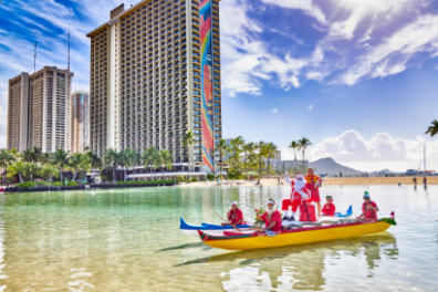 People in red outfits paddling a yellow outrigger canoe near tall buildings on a sunny beach.