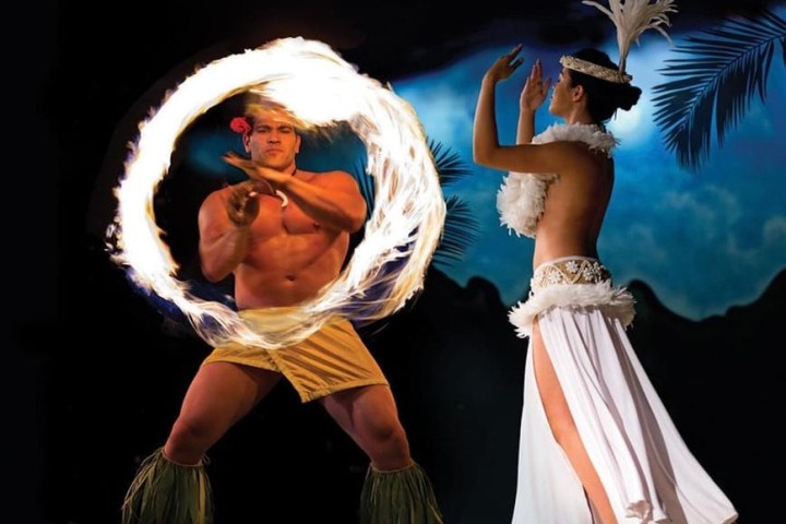 Performers in traditional attire, one spinning fire, the other dancing, with palm silhouettes and a vibrant backdrop.